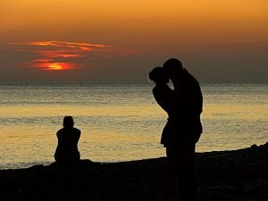 Silhueta de um casal namorando ao pôr-do-sol enquanto uma mulher solitária olha para o mar