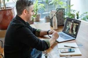 Homem em um sala com algumas plantas, vestindo blusa escura e conversando por vídeochamada no notebook com uma mulher.