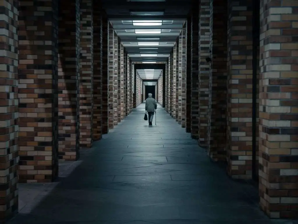 Um homem de costas carregando uma bolsa preta na mão esquerda e uma bengala na mão direita, enquanto caminha por um corredor, e se afastando do observador.