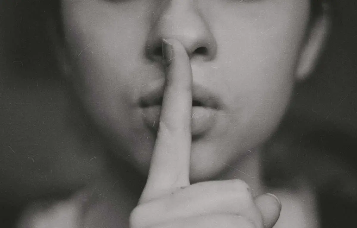 Foto em preto e branco de uma mulher com o dedo em cima da boca, fazendo sinal de silêncio