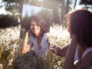 Mulher em um campo de flores, olhando-se em um espelho e sorrindo