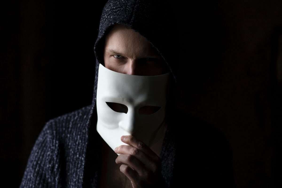 Homem de olhos claros, vestindo um casaco escuro, abaixando uma máscara branca na metade do rosto