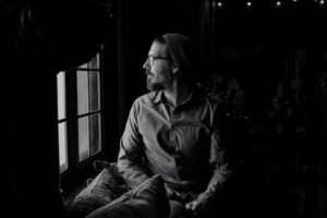 Foto em preto e branco de um homem de barba e óculos olhando pela janela