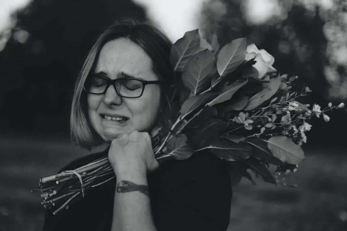Foto em preto e branco de uma mulher chorando, de cabelo curto e óculos, segurando um buque de flores na mão esquerda