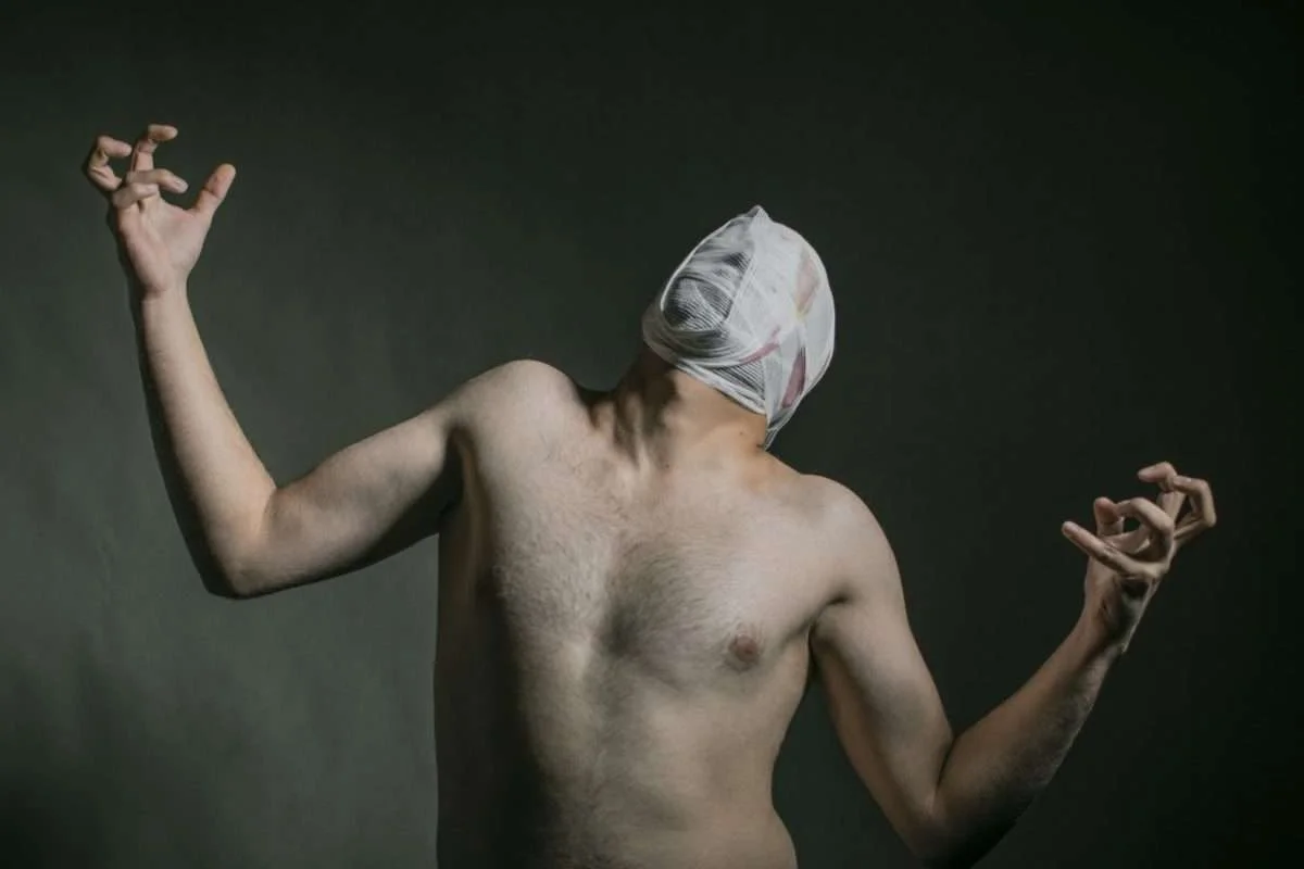Homem sem camisa com faixas em torno da cabeça de modo a esconder o seu rosto, enquanto os dedos das mãos estão dobrados
