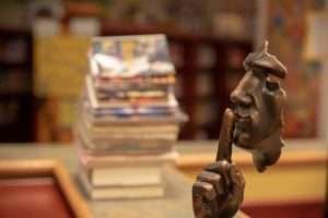 Busto em bronze do rosto de um homem com o dedo na boca, em sinal de silêncio, enquanto ao fundo, desfocado, está uma pilha de livros e revistas