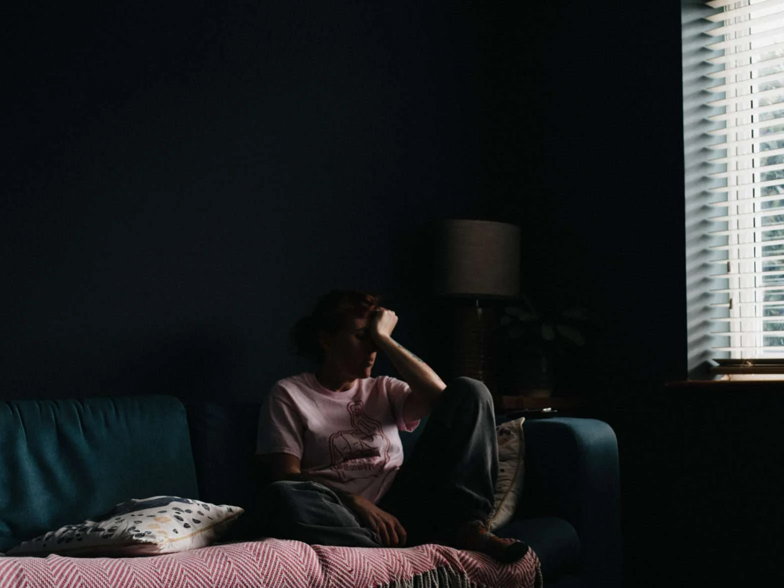 Mulher de camisa rosa sentada em um sofá, de olhos fechados e com a mão no rosto
