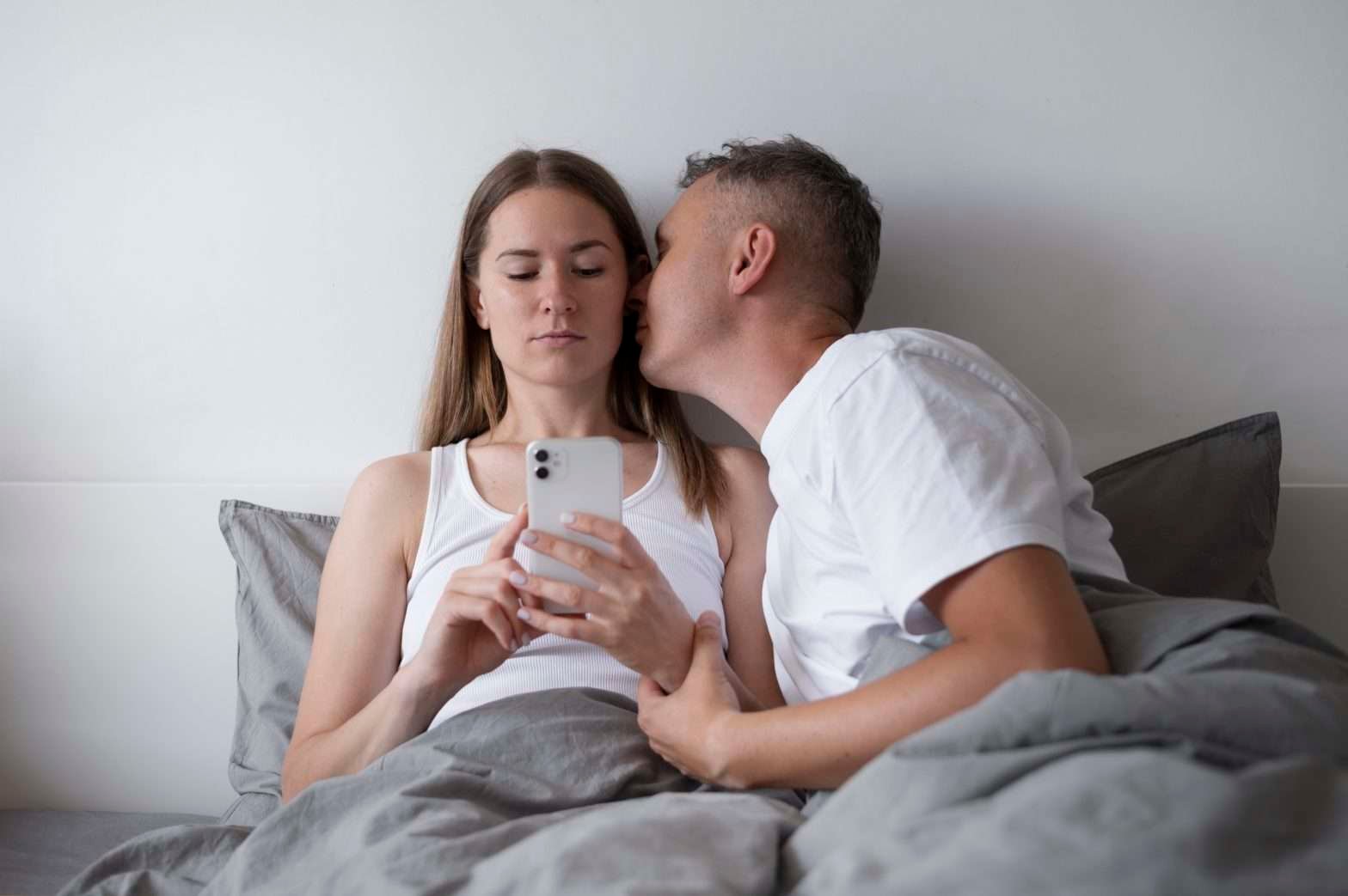 Homem na cama beijando uma mulher no rosto, enquanto ela olha o celular