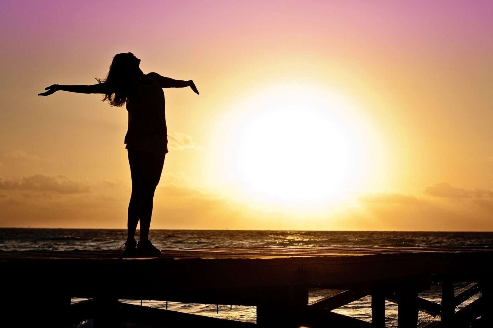 Mulher com os braços abertos em cima de um trapiche durante o nascer do sol