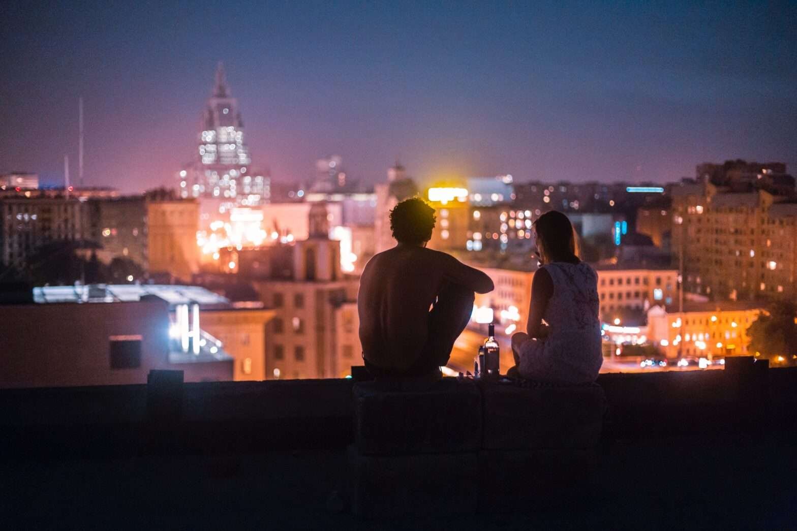 Casal sentado no telhado de um prédio e apreciando as luzes da cidade