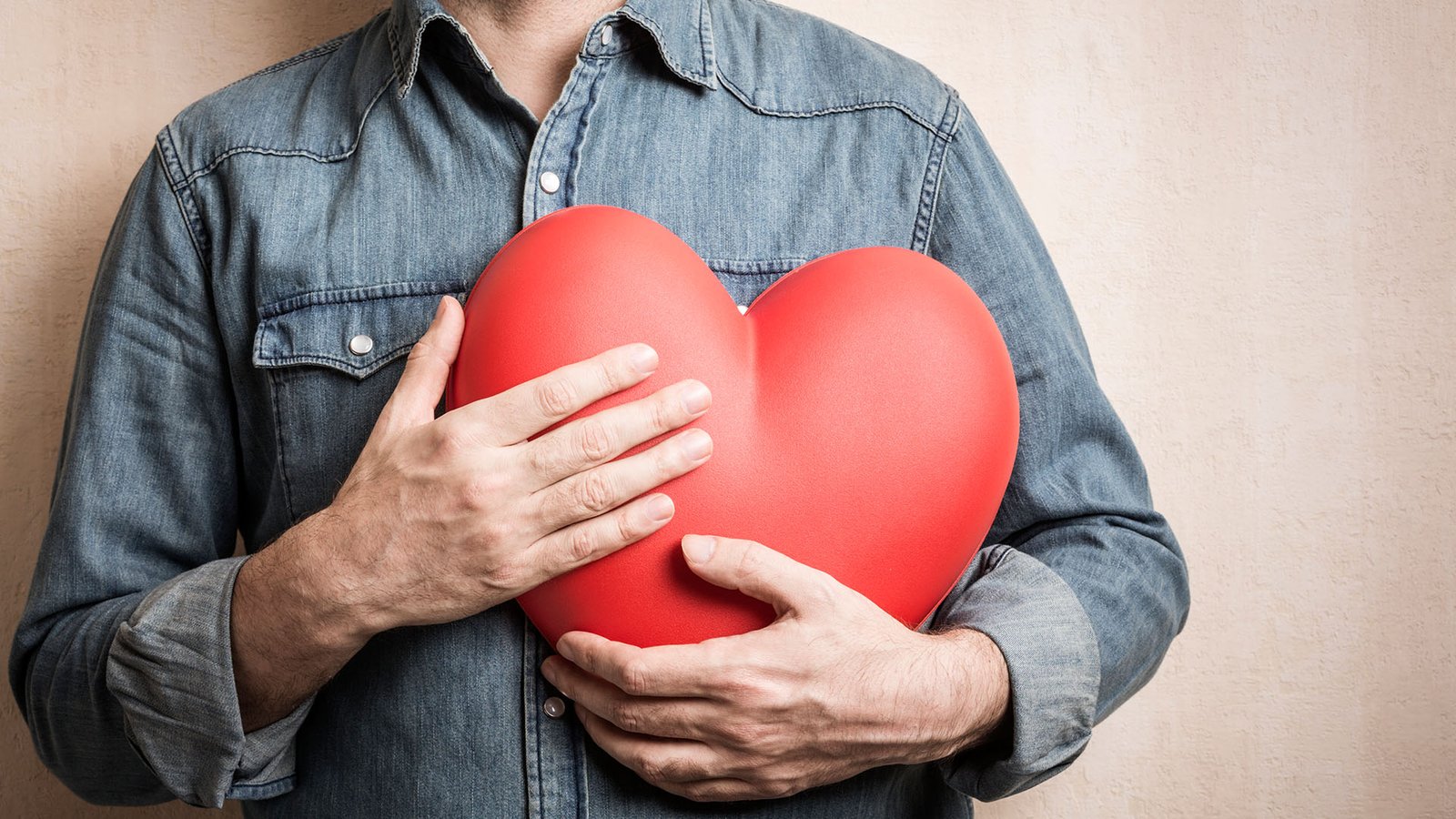 Homem de camisa jeans segurando um coração próximo ao seu peito.