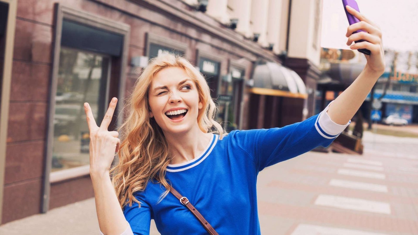 Mulher loira e de camisa azul fazendo uma selfie no meio da rua