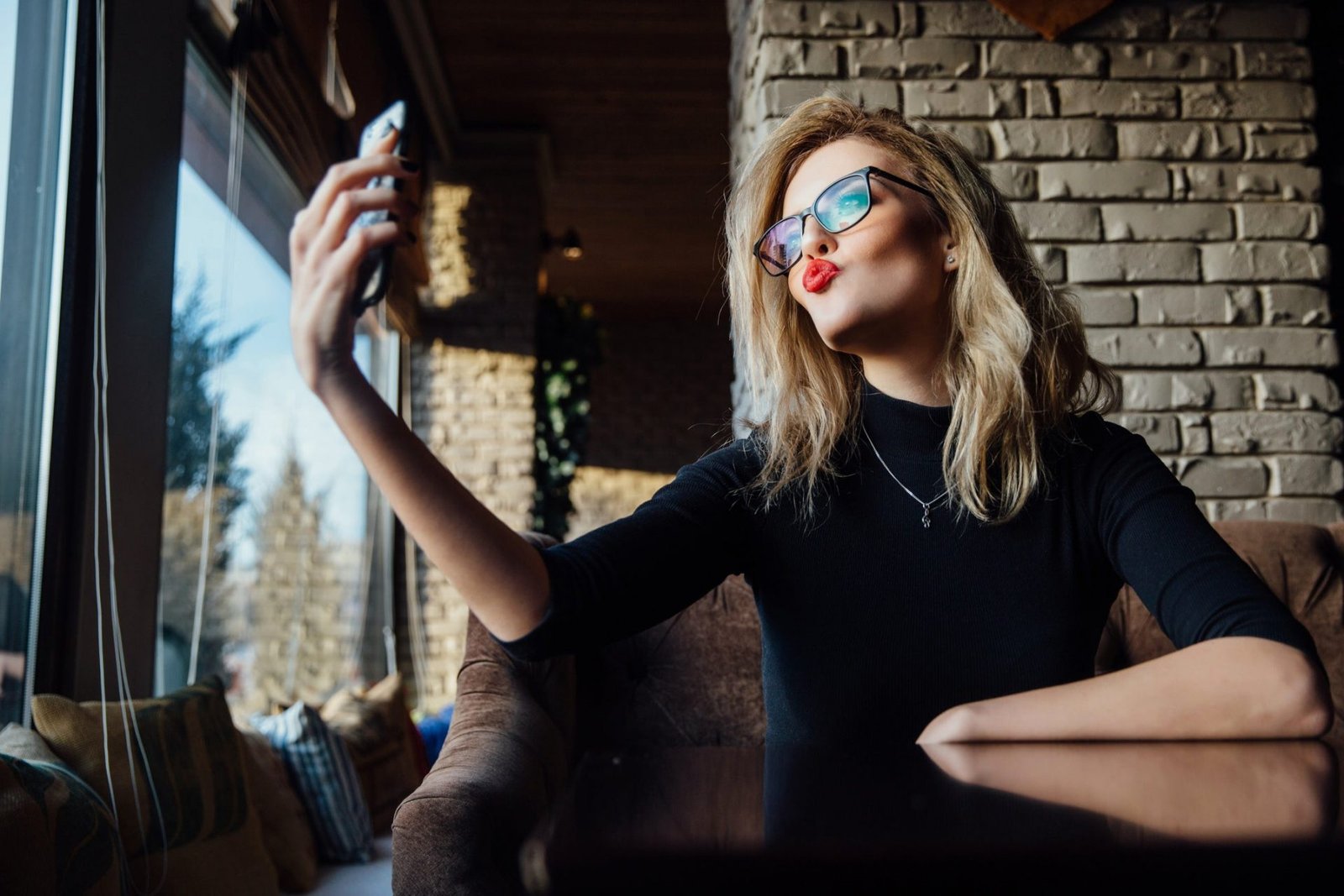 Uma mulher loira, de óculos, bom vermelho e fazendo uma selfie
