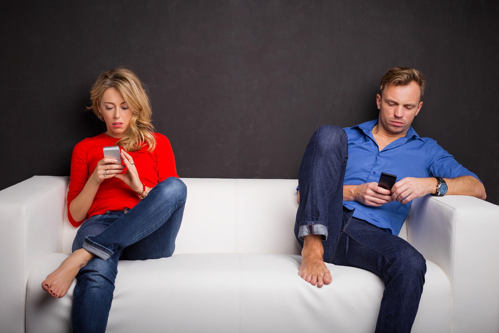 Casal sentado cada um em um canto do sofá, enquanto verificam a tela dos seus celulares.