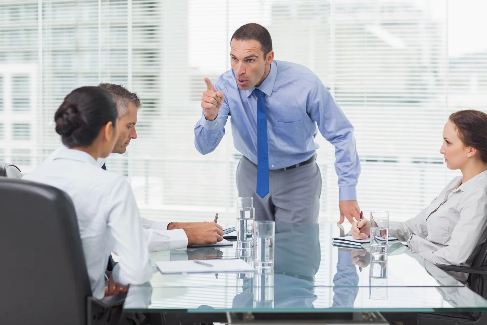 Um chefe narcisista de camisa azul apontando o dedo para colaboradores em uma reunião