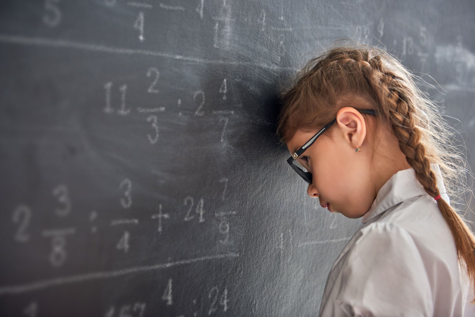 Uma menina de óculos com a cabeça encostada em um quadro negro, com cálculos de frações escritos