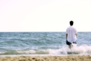Um homem na beira do mar, molhando suas bermudas e camisa com uma onda