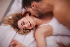 Um casal deitado na cama, onde um homem está por cima da mulher e ela mostrando satisfação